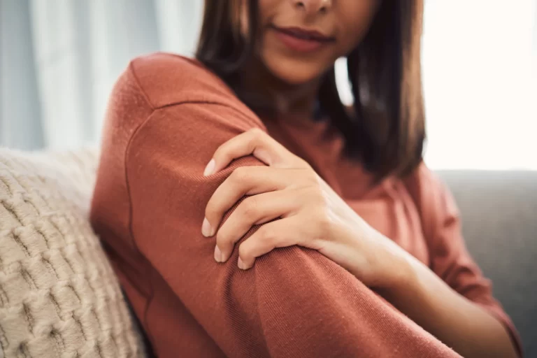After undergoing a brachioplasty procedure, a woman is sitting on a couch with her arm resting on her shoulder, taking part in her recovery and aftercare routine.