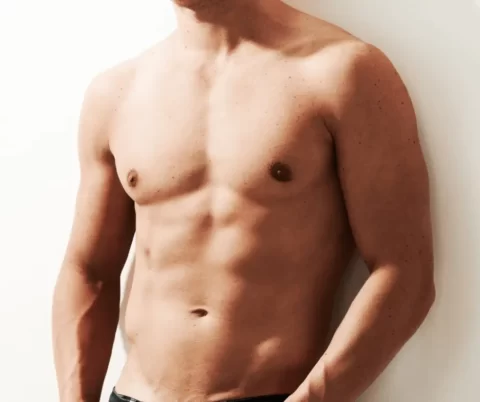 A shirtless man showcasing his defined abdomen as he casually leans against a wall.