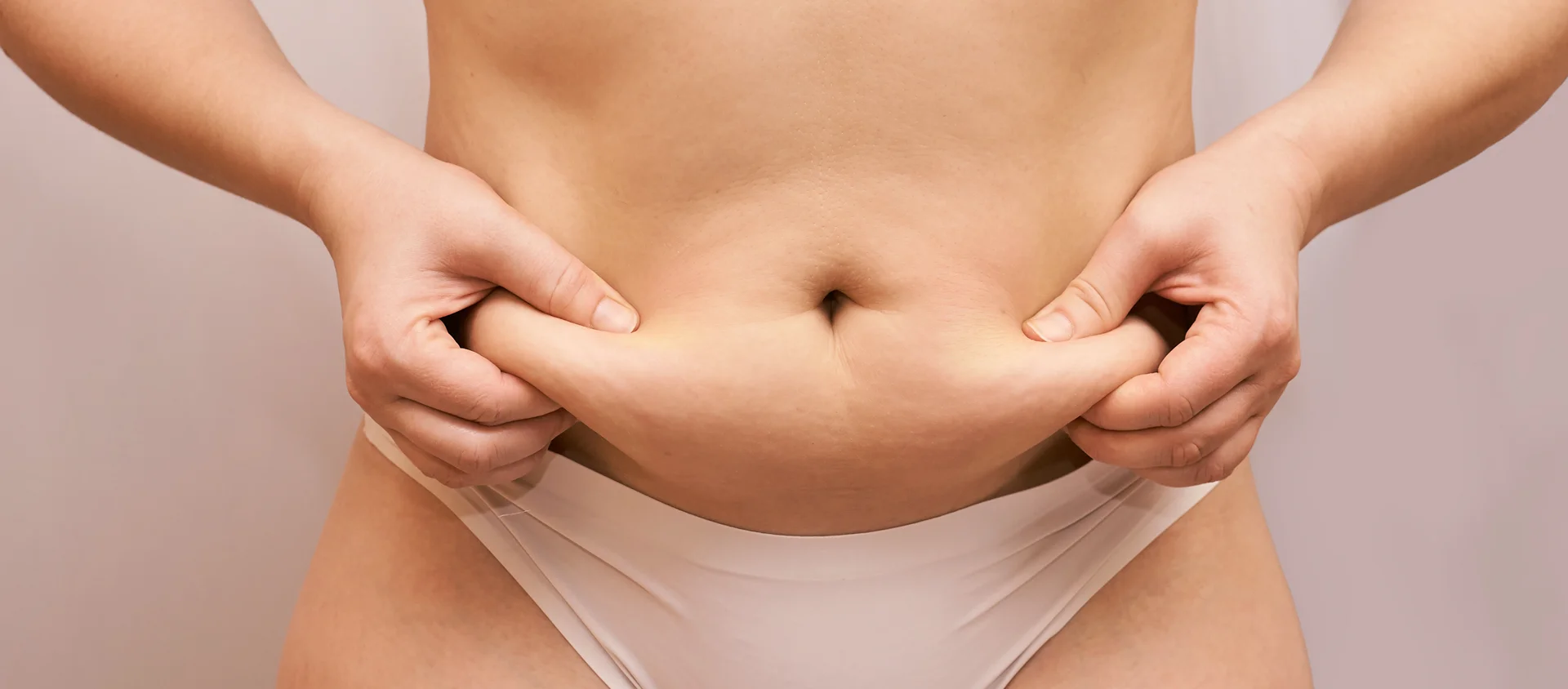 A close-up of a person pinching belly fat while wearing white underwear against a neutral background, indicative of areas targeted by Lipo 360.