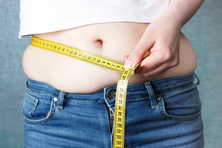 Woman measuring her waist with a yellow measuring tape over blue jeans against a turquoise background after a tummy tuck.