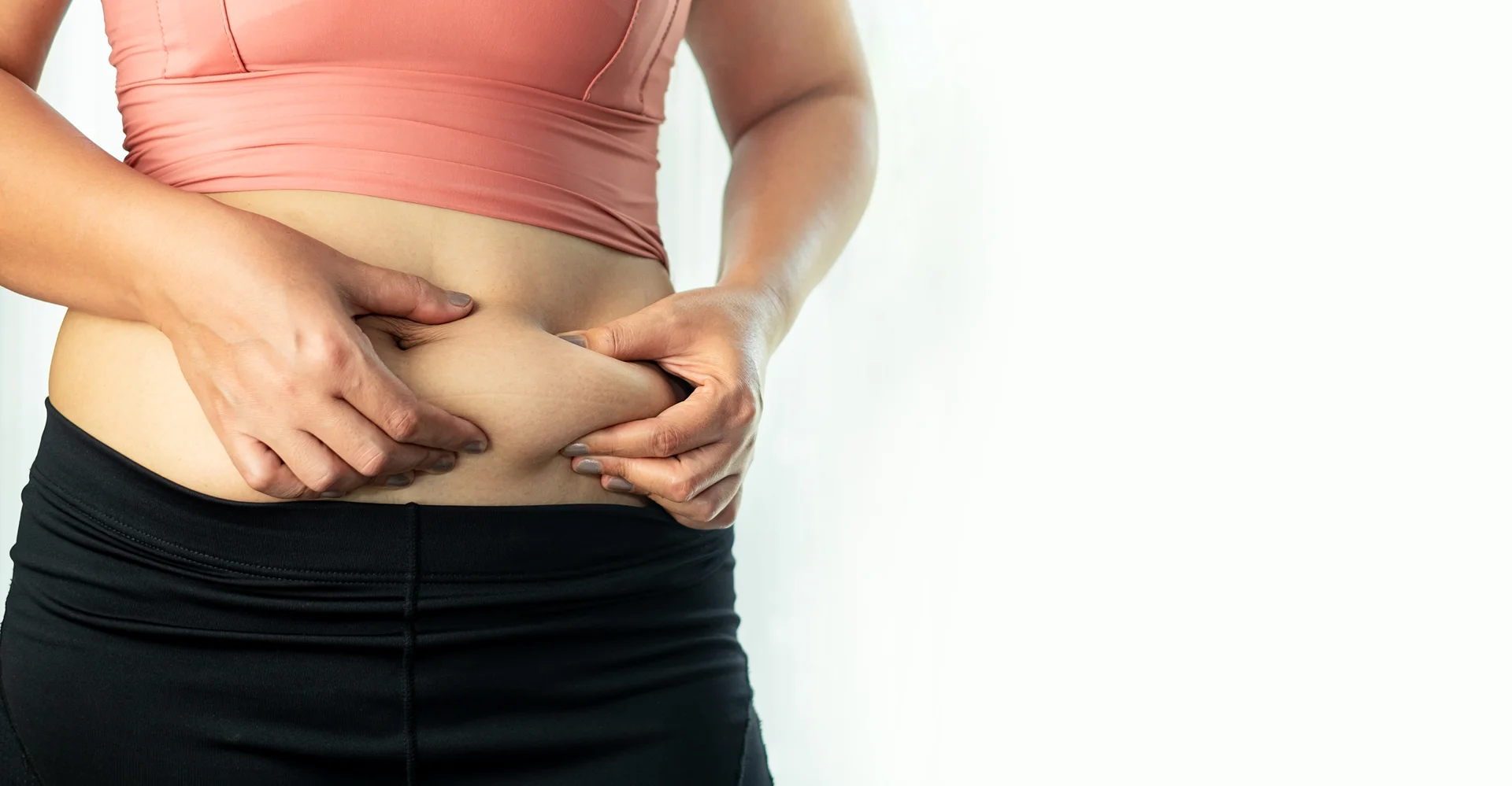Woman pinching belly fat, wearing a pink top and black pants, post Lipo 360 surgery, against a white background.