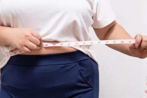 A person wearing a white t-shirt and blue pants, dreaming of their ideal body, measures their waist with a tape measure.