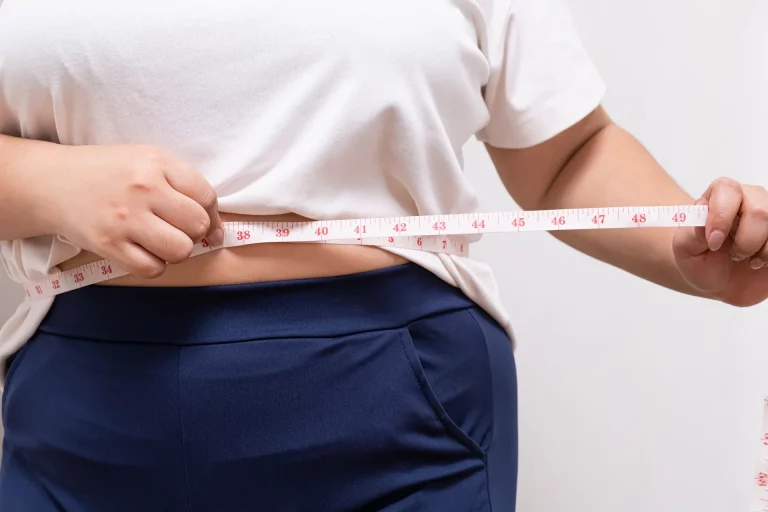 A person wearing a white t-shirt and blue pants, dreaming of their ideal body, measures their waist with a tape measure.