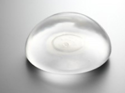 A transparent, dome-shaped silicone breast implant rests on a gray surface.
