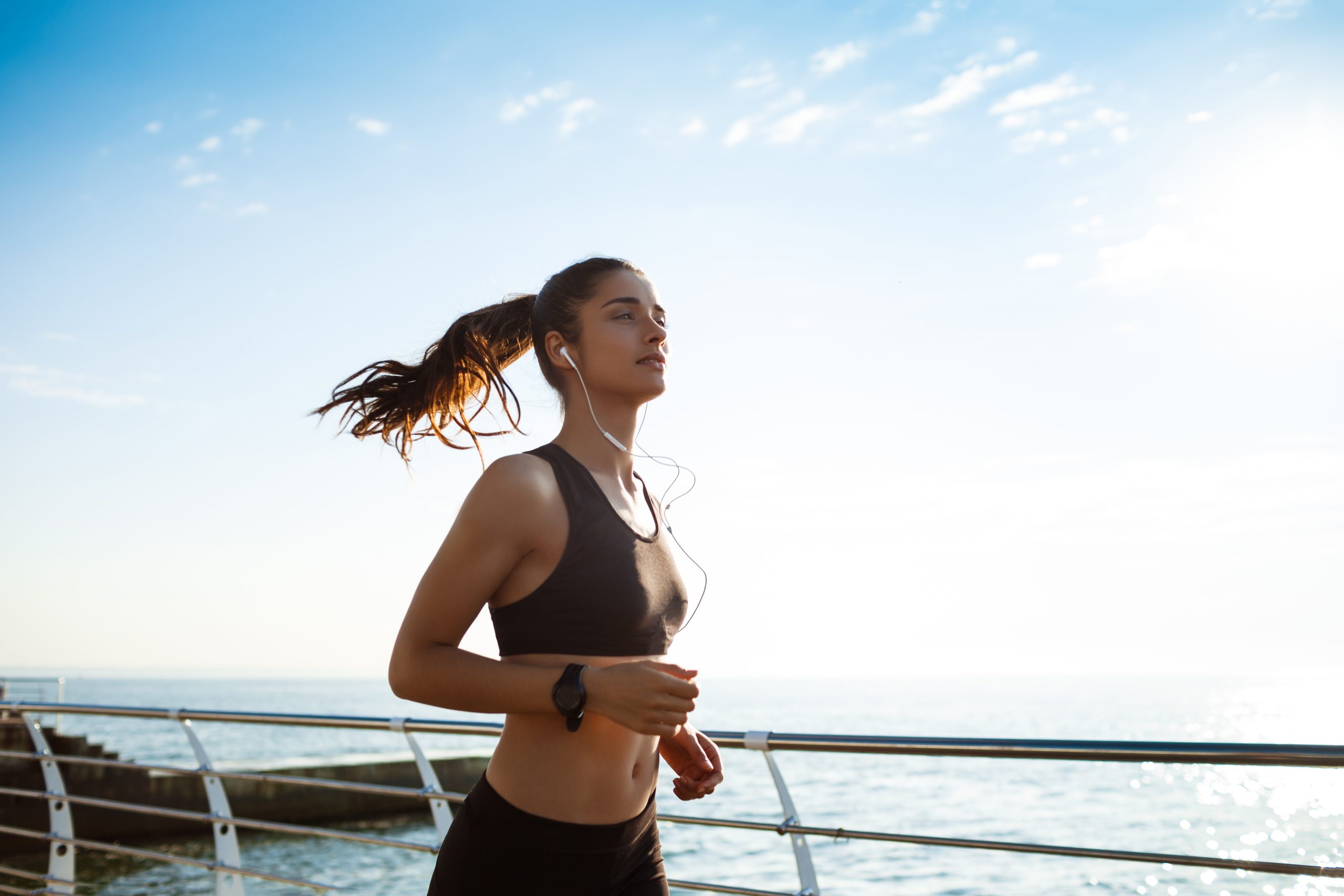 Woman with an athletic body jogs outdoors along a waterfront pathway, wearing a sports bra, fitness tracker, and earphones, with the sea and clear sky in the background.