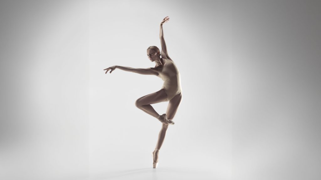 A ballerina in a beige leotard balances on one pointe, with one leg raised and arms gracefully extended against a plain, light background.
