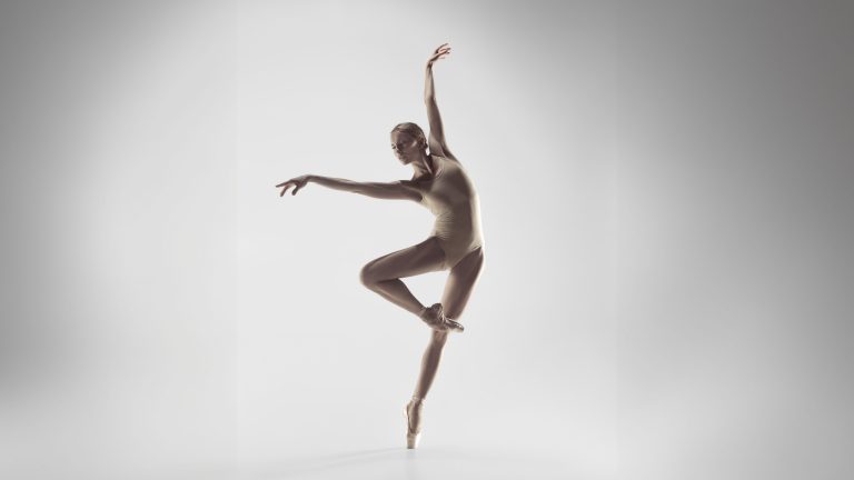 A ballerina in a beige leotard balances on one pointe, with one leg raised and arms gracefully extended against a plain, light background.