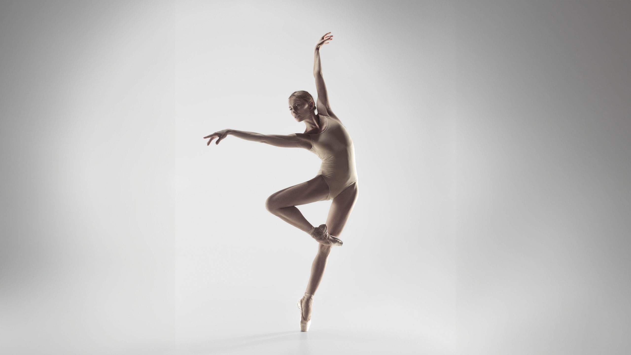 A ballerina in a beige leotard balances on one pointe, with one leg raised and arms gracefully extended against a plain, light background.