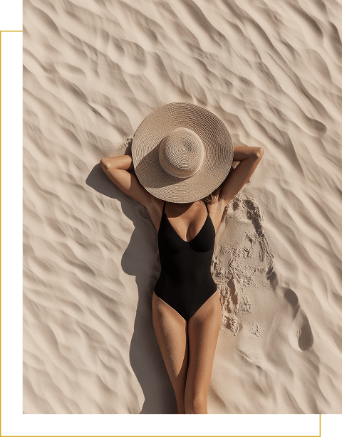 Person in a black swimsuit lies on sandy terrain with arms behind their head, face covered by a wide-brimmed straw hat.