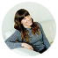 A woman with long brown hair and bangs, wearing a gray sweater, sits on a white couch and smiles at the camera.
