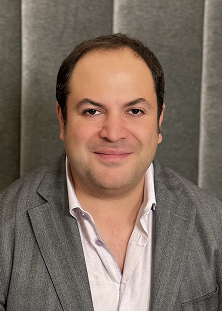 A man with short dark hair wearing a light pink shirt and a grey blazer sits in front of a gray padded background, smiling at the camera.
