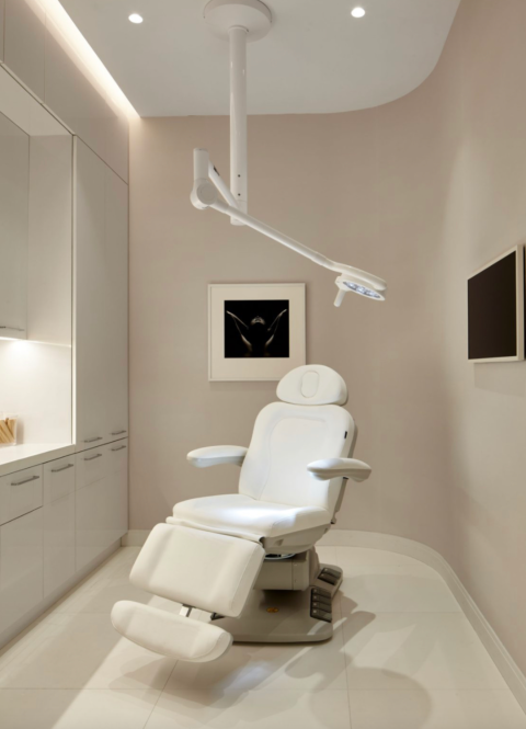 Modern medical examination room with a white adjustable chair, overhead light, cabinets, wall art, and a mounted screen.