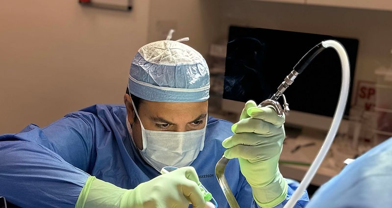 Dr. Darren Smith, dressed in blue scrubs and gloves, performs a medical procedure using specialized equipment in a clinical setting.