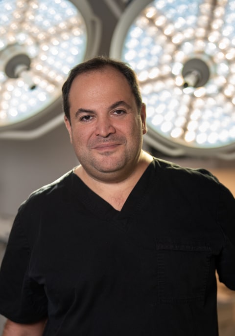 Dr. Darren Smith, wearing black medical scrubs, stands in front of two large surgical lights in what appears to be an operating room.