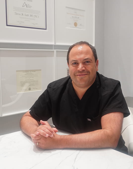A man in black scrubs sits at a marble table with his hands folded, smiling. Behind him, framed certificates and diplomas are displayed on the wall, suggesting confidence and expertise—perfect for discussing your new home layout plans for 2026.