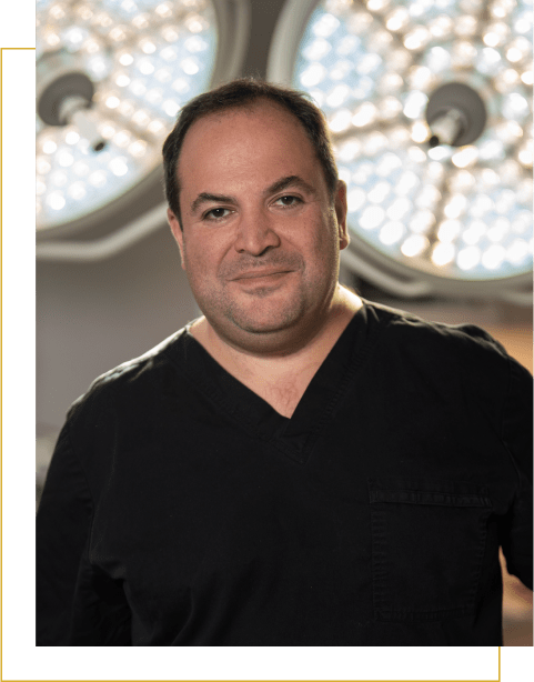 Dr. Darren Smith, dressed in black medical scrubs, stands in front of large, round surgical lights, facing the camera and smiling slightly.