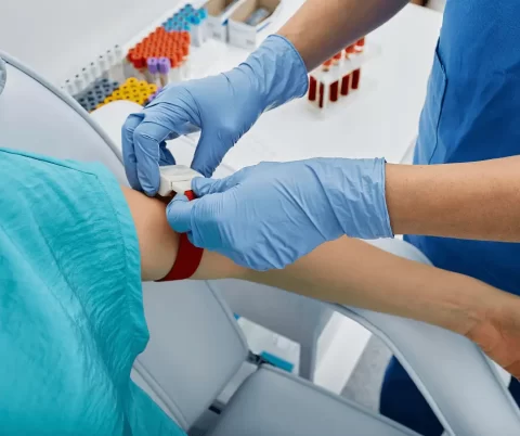 A healthcare worker wearing blue gloves prepares a patient's arm for blood draw, with blood sample tubes visible in the background.