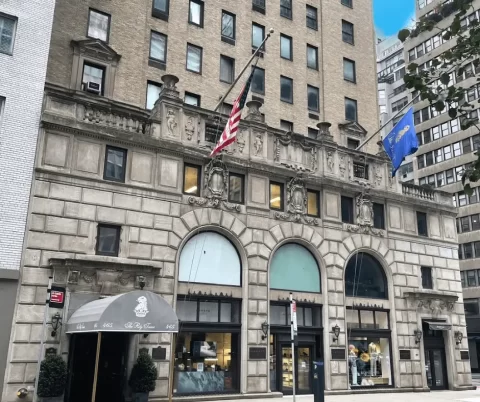 Historic stone building with arched windows, American and blue flags, awning over entrance, and tall modern buildings in the background.