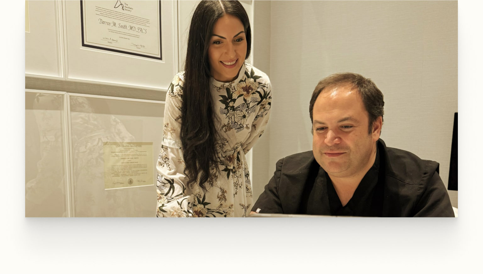 A woman in a floral dress stands next to a seated man in dark clothing as they look at a computer, with framed certificates on the wall behind them.