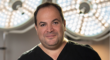 A man in black scrubs stands in front of bright surgical lights, looking at the camera with a neutral expression.