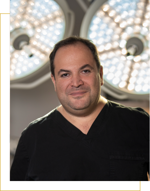 A man in black scrubs stands in front of two large surgical lights, smiling at the camera, ready for a Plastic Surgery procedure.