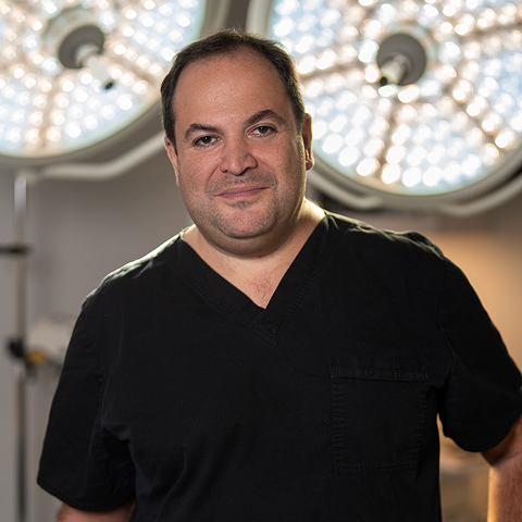A man in black medical scrubs stands in front of round surgical lights in a clinical setting, ready to assist both local and Out of Town Patients.