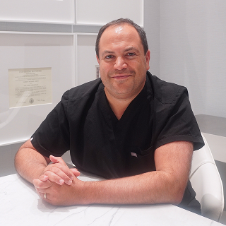 A man in black medical scrubs sits at a marble desk with hands clasped, smiling, in a modern office with a diploma on the wall behind him—ready to help you. Schedule a consultation today for expert care.