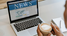 A person holds a cup of coffee while viewing an online news website on a laptop, with a notepad and pen nearby.