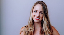 A woman with long blonde hair and a bright smile is standing in front of a plain light-colored background.