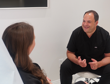 A man in scrubs sits and talks with a woman in a medical office, both appearing engaged in conversation about her care at the practice.
