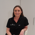 A woman with long brown hair, wearing a black practice uniform, sits on a white chair, smiling, in a modern, light-colored room.