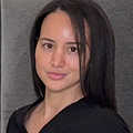 A woman with straight dark hair and a neutral expression poses in front of a plain gray background, as if in quiet practice for a portrait session.