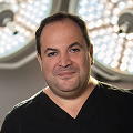 A man with short dark hair wearing a black shirt stands indoors, where blurred round lights in the background evoke the calm focus of practice.