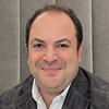 A man with short dark hair, wearing a gray blazer and light shirt, smiles at the camera in front of a gray upholstered background, ready to share his reviews and testimonials.