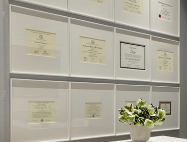 Nine framed certificates are arranged in a grid on a wall above a table, showcasing the achievements of the practice, with a small bouquet of white flowers in a vase adding an elegant touch.