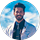 A man with dark hair and a beard, wearing a light-colored blazer and black shirt with a tie, stands in front of a sky background, appearing ready to share reviews or testimonials.