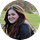 A person with long brown hair, wearing a dark top, is sitting outdoors on grass with trees and people in the background, appearing relaxed as if reading reviews or sharing testimonials.