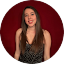 A woman with long brown hair wearing a sleeveless patterned top stands in front of a plain red background, smiling at the camera, as if ready to share her testimonials or reviews.