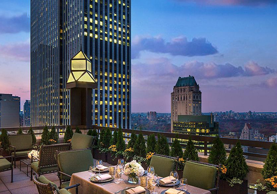 Outdoor dining area on a terrace overlooking city skyscrapers and a park at sunset, with tables set and greenery along the railing—perfect for out of town patients seeking relaxation after a long day.