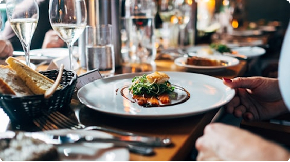 A plate of gourmet food sits on a wooden table set with bread, glasses of white wine, and water at a busy restaurant, welcoming out of town patients looking for a memorable dining experience.