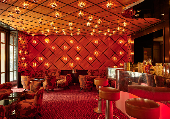 A retro-style lounge with red patterned walls, star-shaped ceiling lights, plush armchairs, and a mirrored bar with gold accents—perfect for out of town patients seeking comfort and style during their visit.