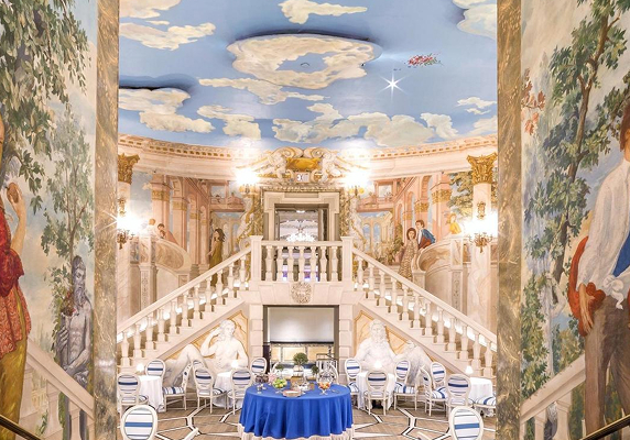 Elegant ballroom with a double staircase, detailed wall murals, blue sky ceiling art, and seating for out of town patients at round tables draped in blue tablecloths, with white chairs arranged throughout the room.
