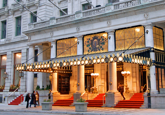 Entrance of a grand hotel with a marquee, red carpeted steps, ornate lights, and people walking by on a city street—ideal for welcoming out of town patients seeking comfort and luxury during their stay.