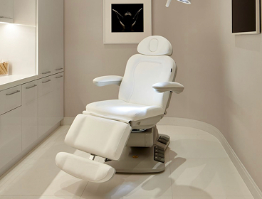 A modern white medical examination chair is positioned in a clean, well-lit clinic room, showcasing a professional practice with organized cabinets and advanced medical equipment.