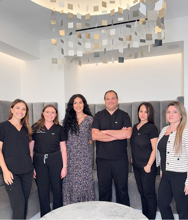 Six people stand indoors, five wearing medical scrubs and one in a dress, posing in front of a modern light fixture and gray cushioned wall, reflecting their teamwork in a professional practice.