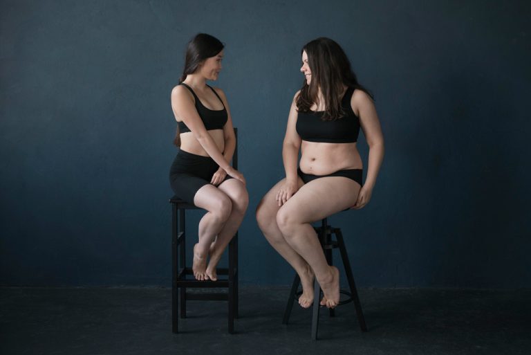 Two women in black underwear sit on stools facing each other, smiling, against a plain dark blue background—celebrating their differences and raising awareness for Lipedema and Lymphedema.